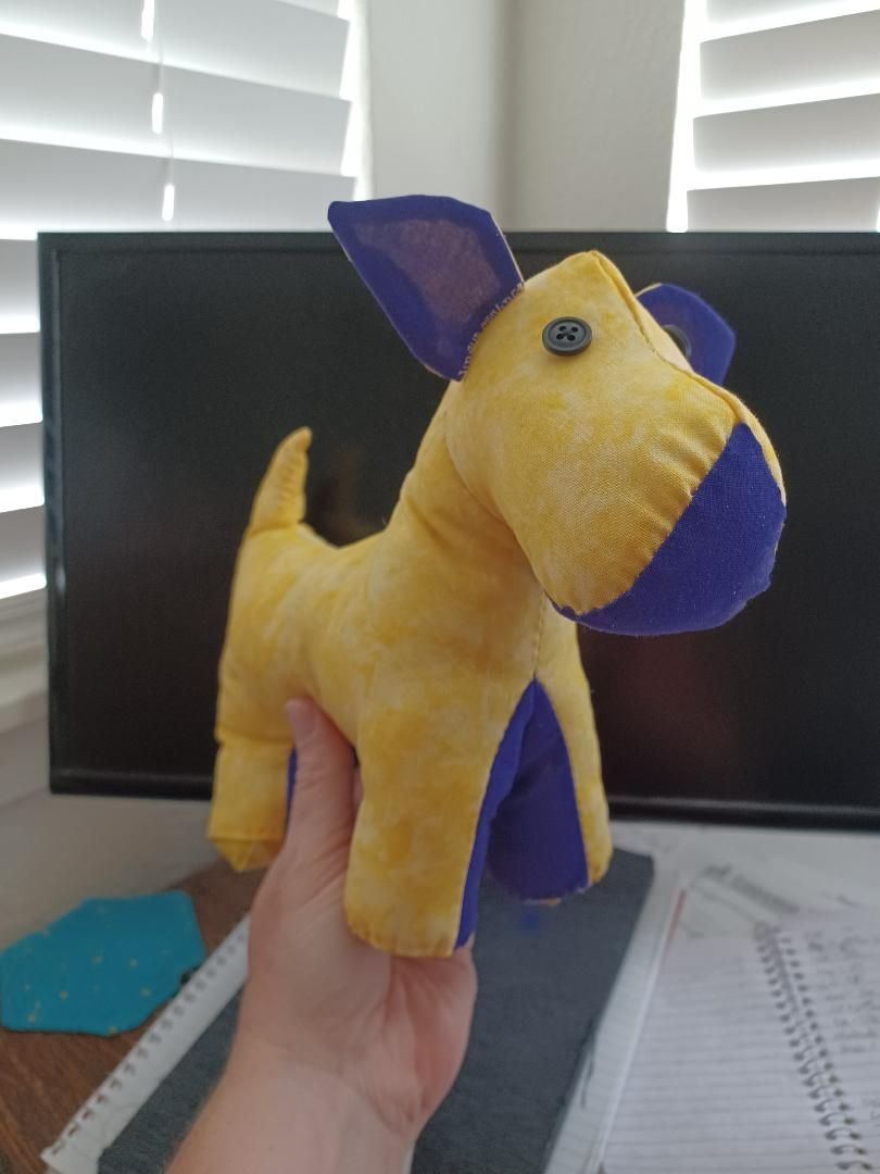 Yellow plush dog toy with blue ears and collar, held up in a hand in front of a computer monitor.