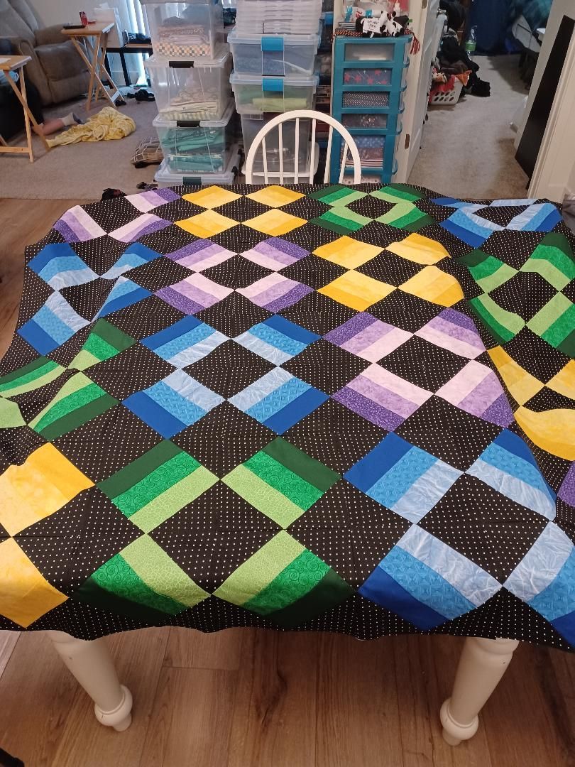 Colorful patchwork quilt draped over a table in a cluttered room with storage bins and a chair behind it