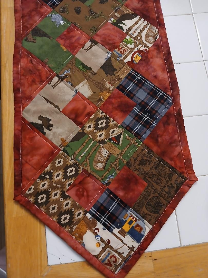 Patchwork quilt runner in red, green, brown, and plaid squares hanging vertically on a wall.