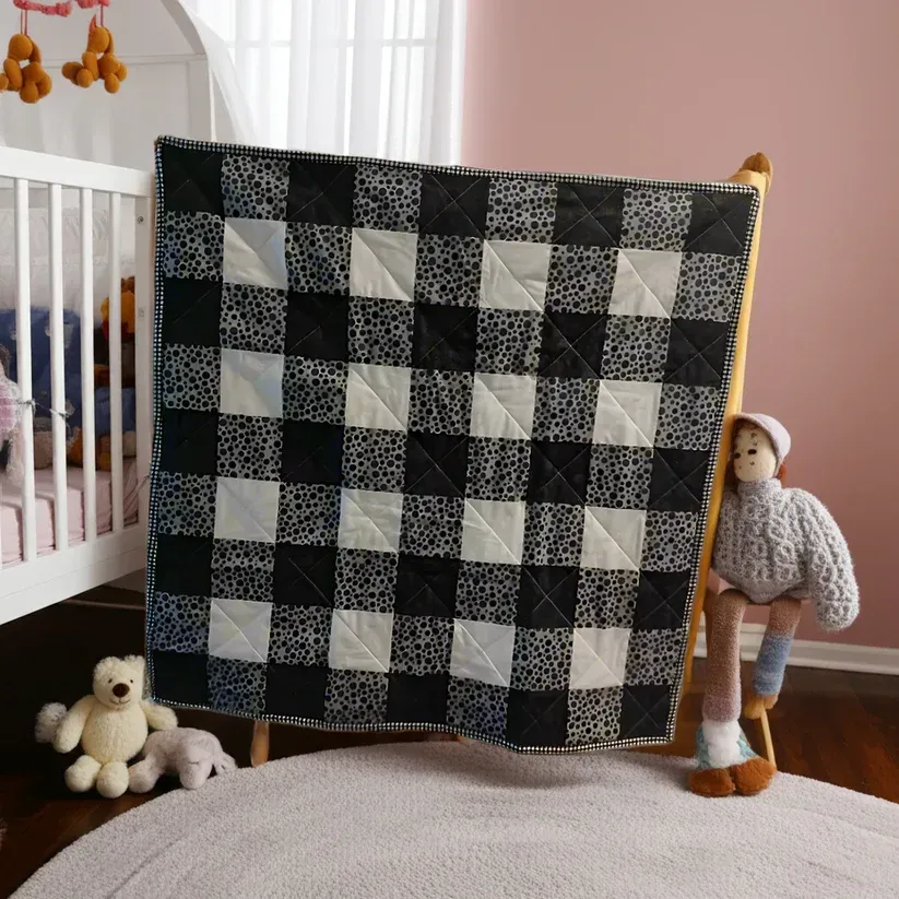 Black-and-white patchwork quilt draped over a crib in a nursery with stuffed toys and a doll nearby