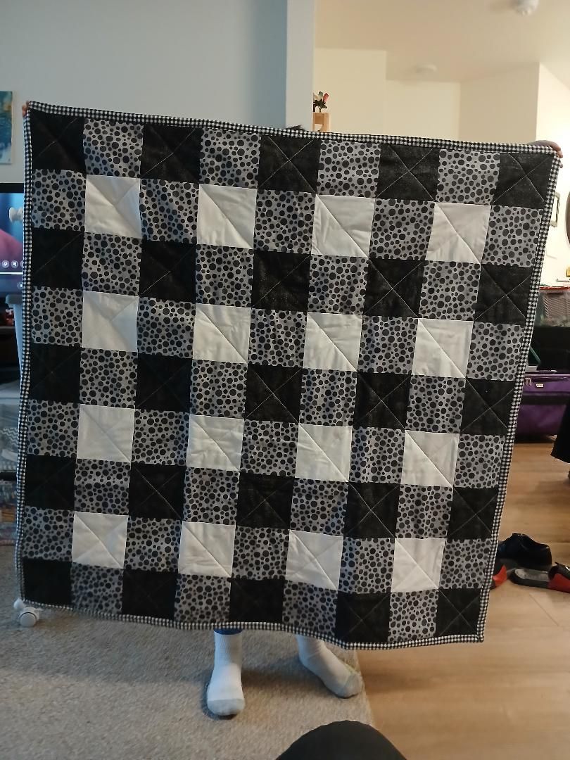 Person holding a large black-and-white checkered blanket indoors; only legs and feet visible.