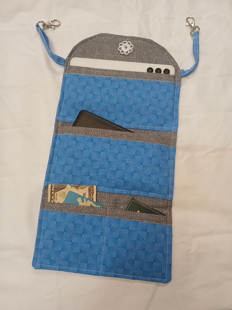 Blue woven pouch with gray top flap, silver snap, and shoulder straps hanging on a wall