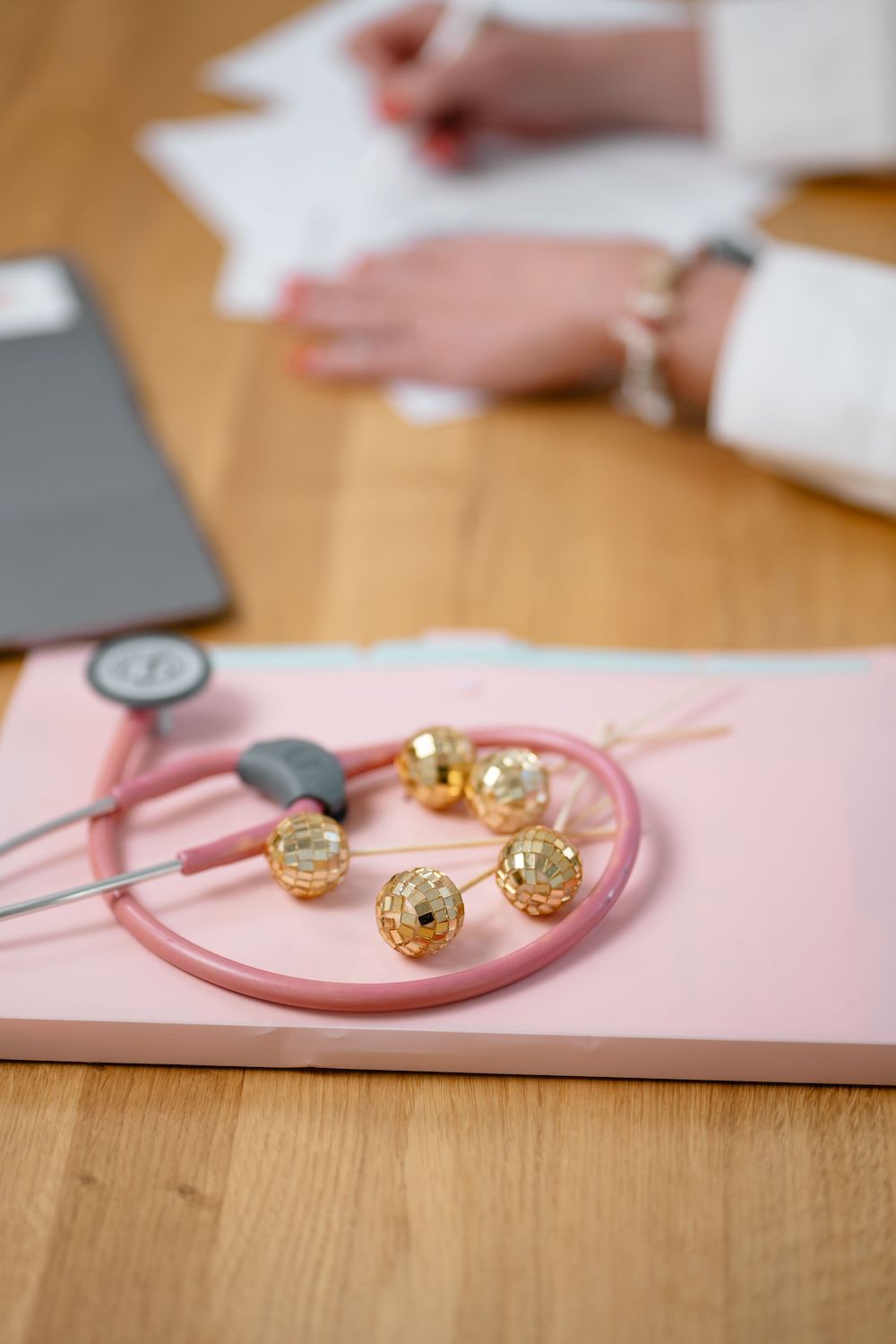 Rosa Stethoskop und goldene Perlen auf einem rosa Block, im Hintergrund eine schreibende Person.
