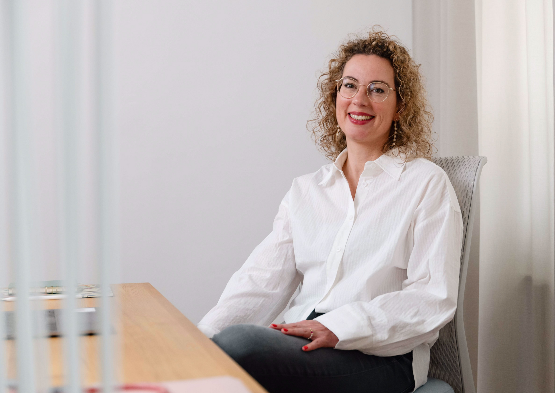 Frau mit lockigem Haar, Brille, weißem Hemd und lächelnd, sitzt an einem Schreibtisch in einem Raum.