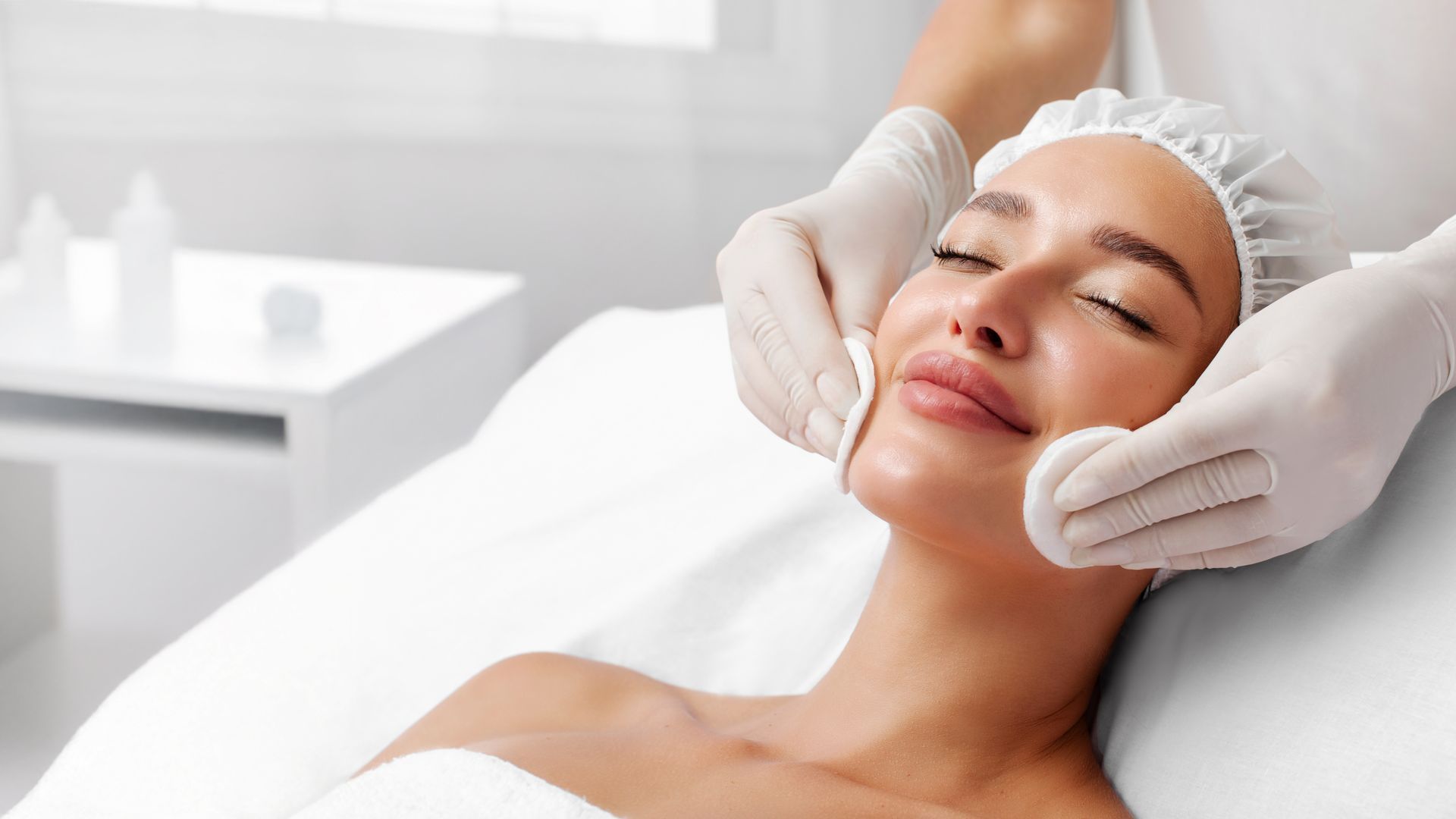 Woman receiving facial treatment; esthetician using device on her forehead. Indoors, white and pink colors.