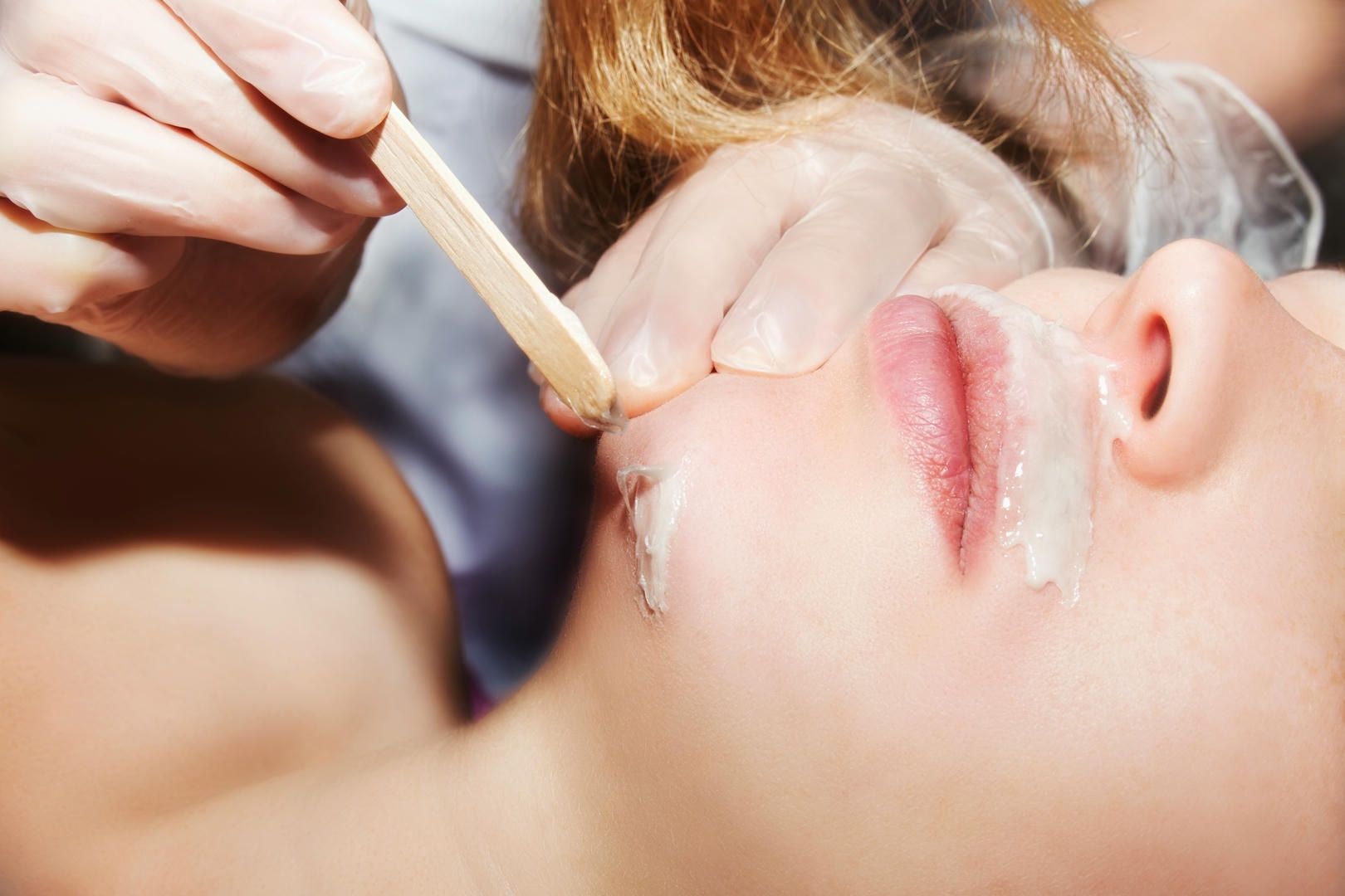 Person having upper lip waxed; wax applied by gloved hands with wooden spatula.