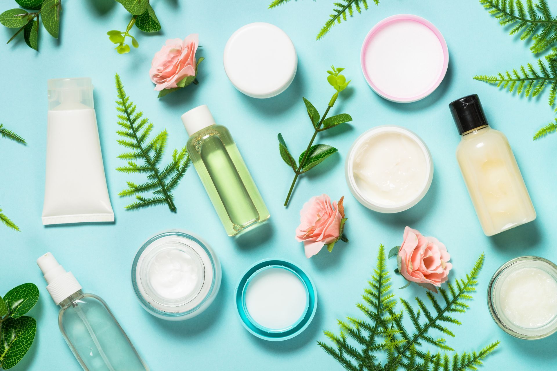 Skincare products, pink roses, and fern leaves arranged on a light blue surface.