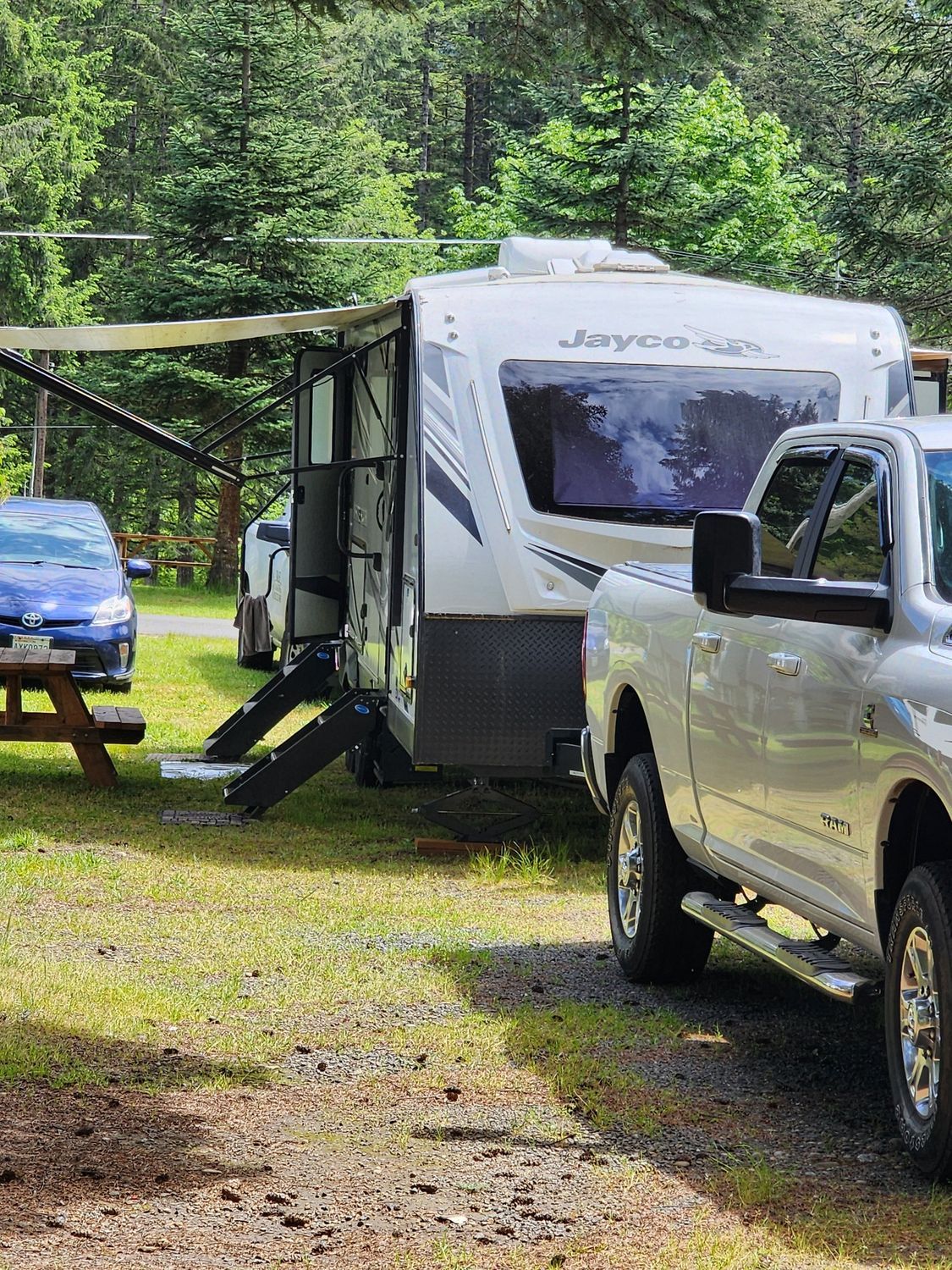 White camper trailer and pickup truck parked on grass beside a blue car in a wooded campsite.