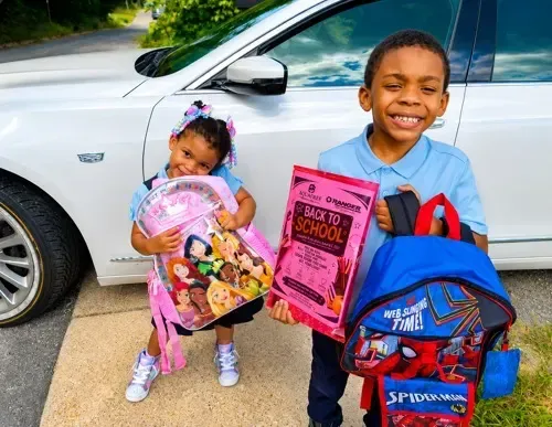 Two smiling children with backpacks and school supplies standing by a silver car