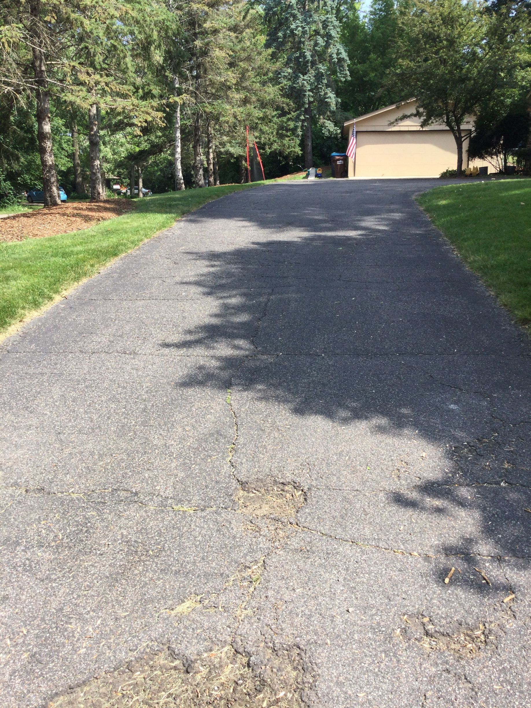 Asphalt driveway with cracks, leading to a garage, surrounded by grass and trees.
