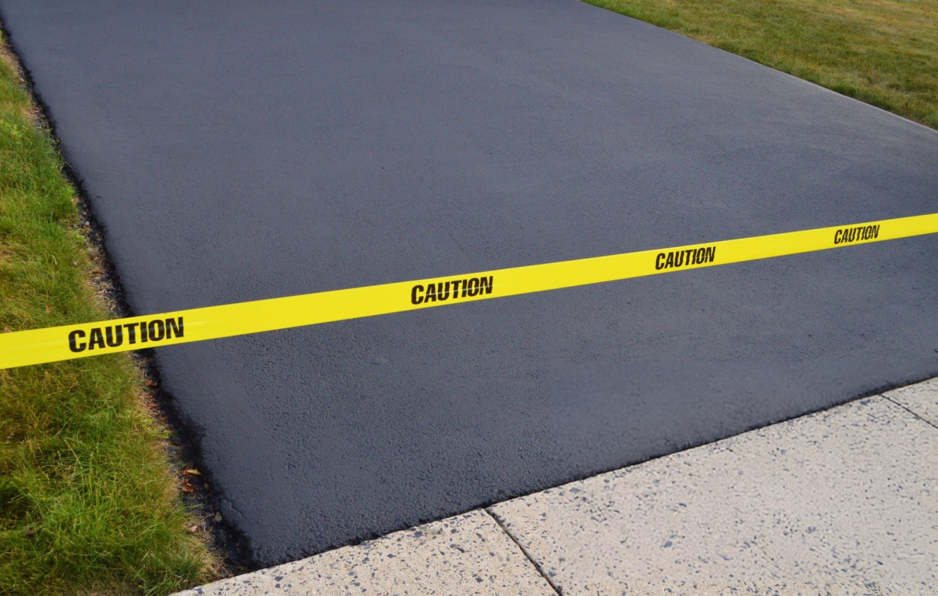 A newly seal-coated asphalt driveway against a sidewalk A newly seal-coated asphalt driveway against a sidewalk