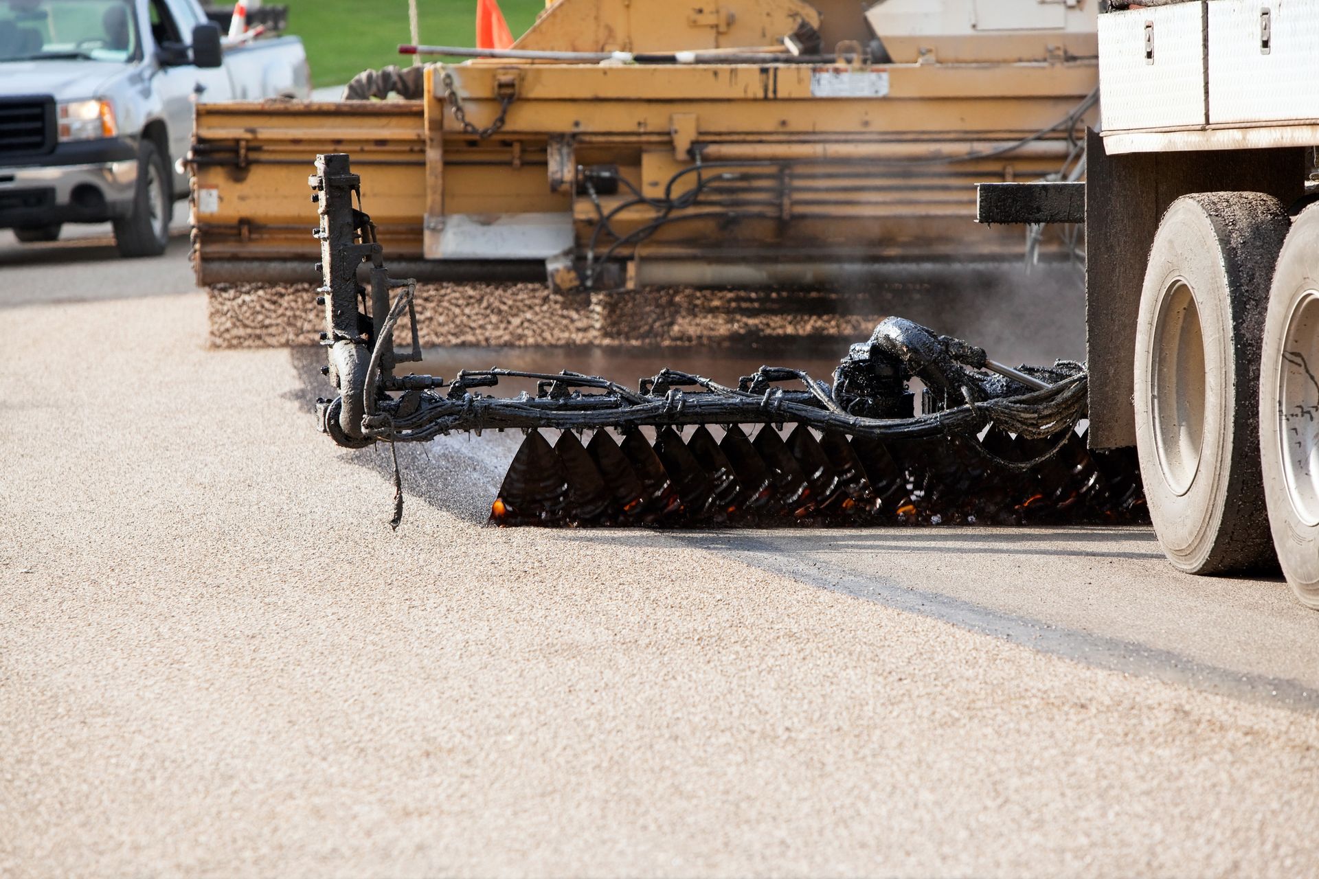 Black asphalt sealcoat is being applied on the road and white truck behind.