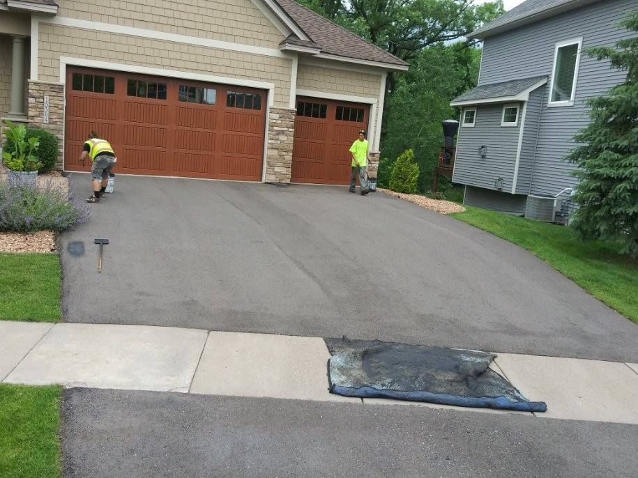 Two workers seal a black asphalt driveway in front of a house with brown garage doors.