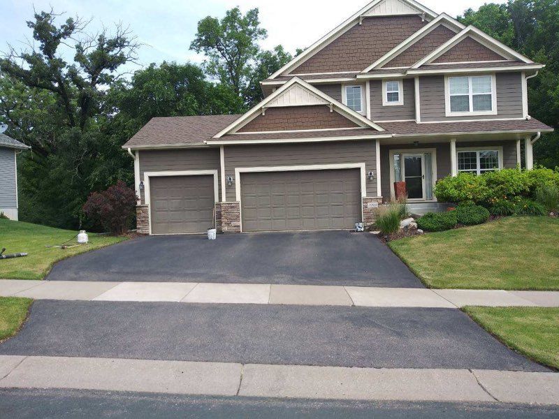 Brown house with a two-car garage and a long asphalt driveway, surrounded by green grass and trees.