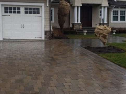 Brick driveway leads to a white garage door, house front. Newly planted trees wrapped. Wet.