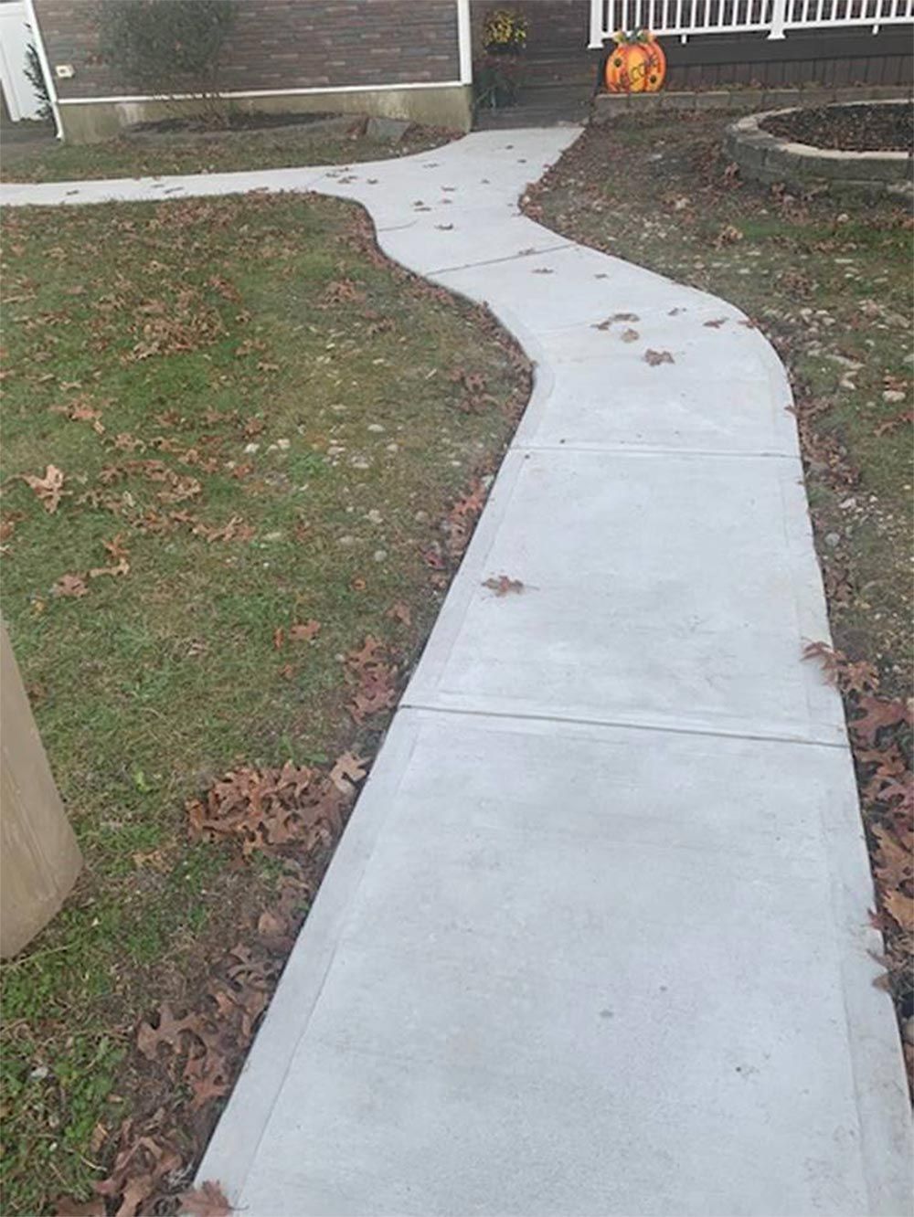 A winding concrete sidewalk leads to a house with a pumpkin on the porch, surrounded by grass and fallen leaves.