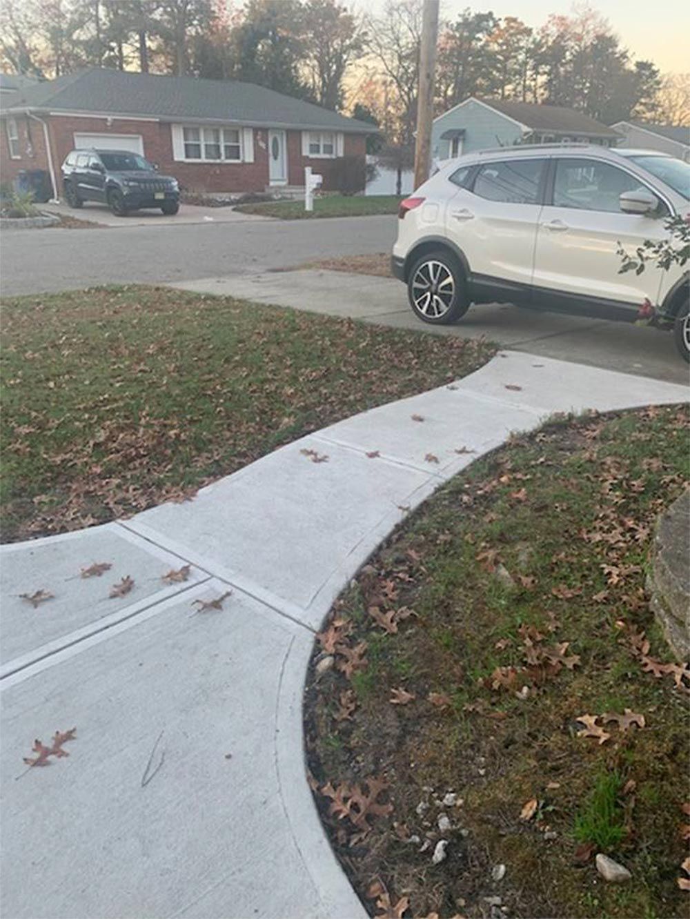 Concrete sidewalk curves along a lawn with fallen leaves, a white SUV parked nearby, and houses in the background.