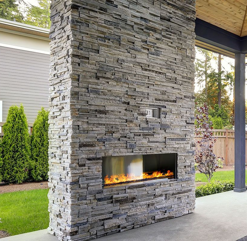 Stone fireplace with flames, built into a covered patio, in front of a green lawn and trees.