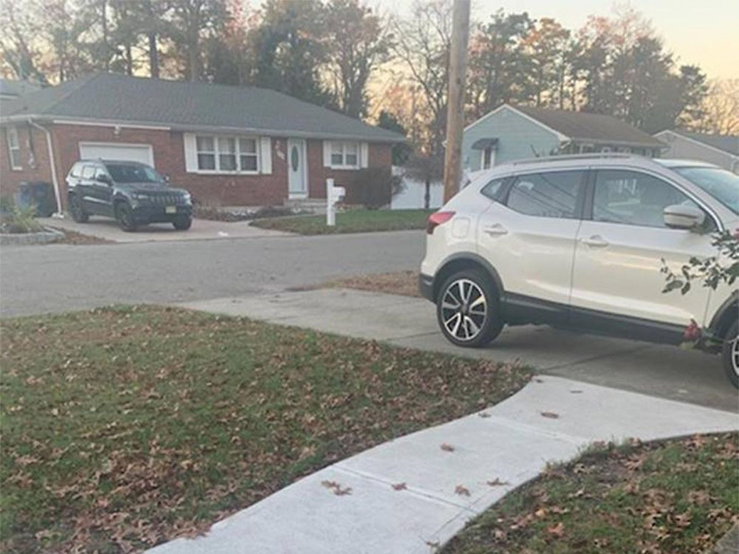 A white SUV parked in a driveway, in front of a brick house with a dark SUV parked in front of it.