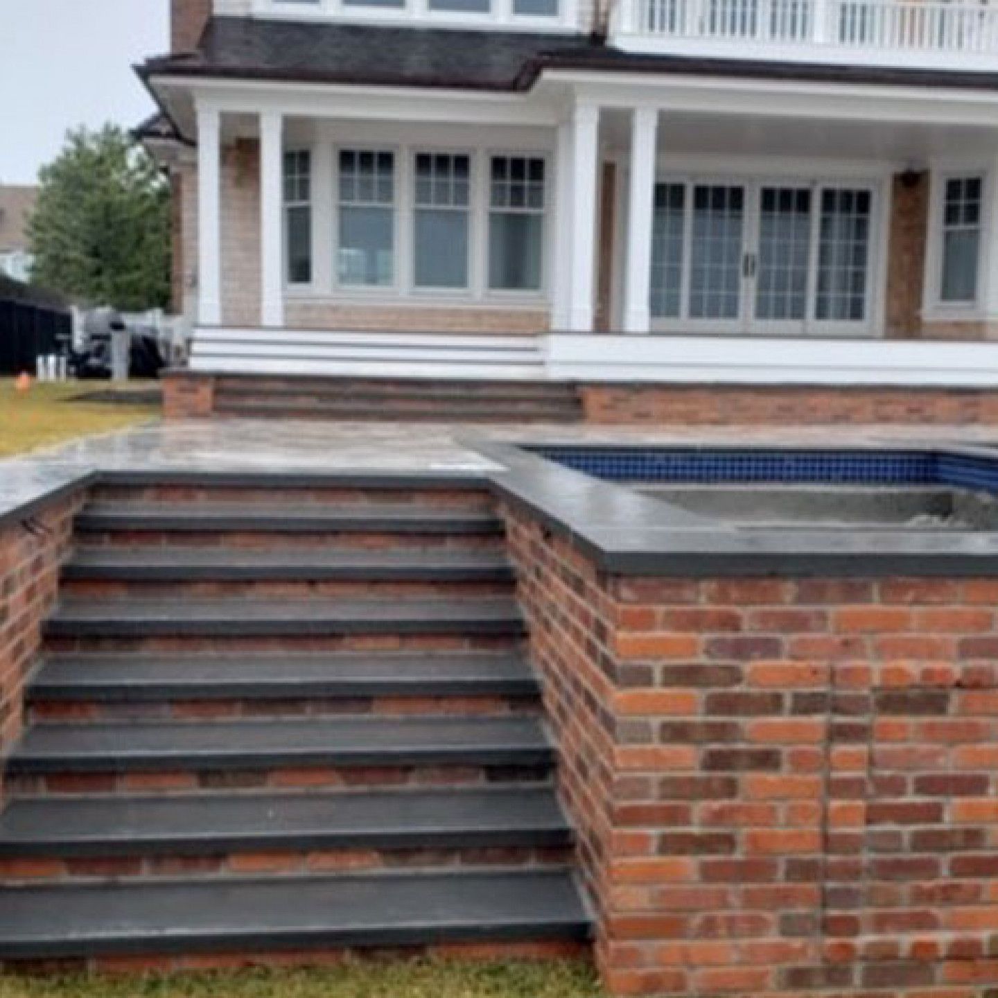 Brick staircase leading up to a patio with a swimming pool, attached to a house with a white porch.