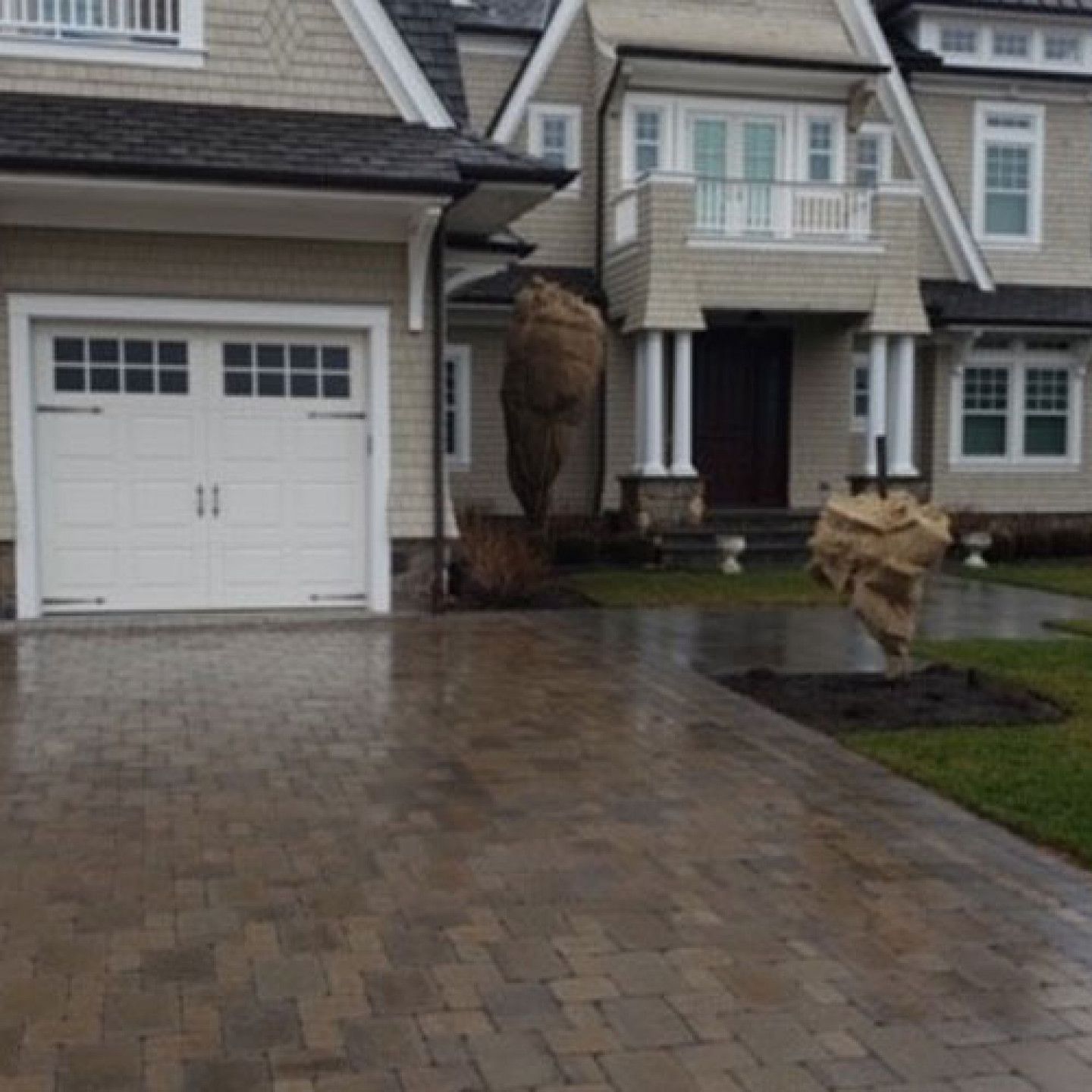 Paver driveway leading to a beige house with a garage and covered porch. Two wrapped trees are in the front yard.