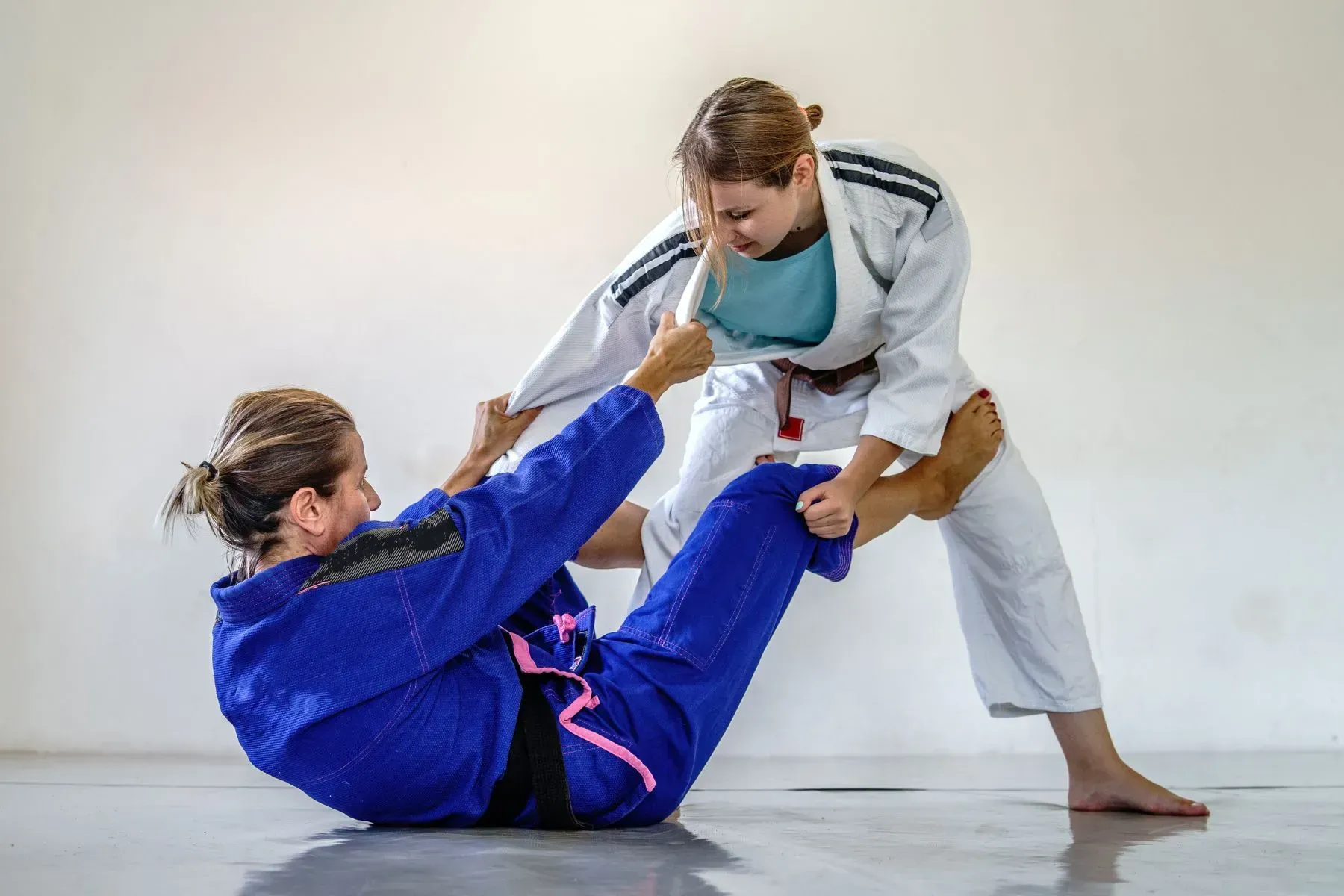 Two judo practitioners in white and blue gis grappling on a mat, one seated and one standing.