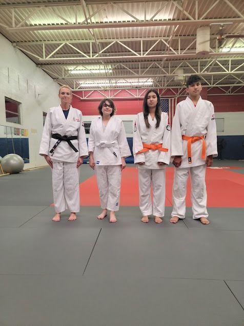 Four martial arts students in white uniforms stand on a gym mat in an indoor training hall.