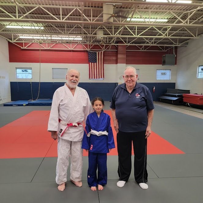 Three people standing on a red martial arts mat in a gym, with an American flag in the background