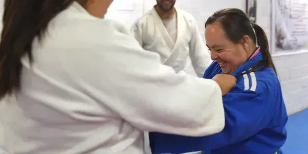 Two people in judo gis practicing a hold, one in blue smiling in a training room