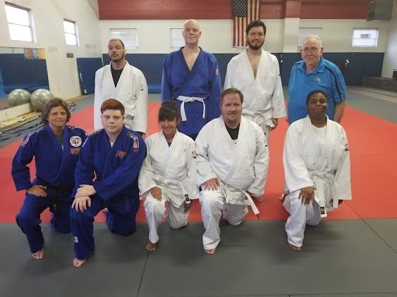 Group of judo practitioners posing on a red mat in a dojo, wearing blue and white gis.