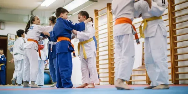 Children practicing judo in white and blue uniforms in a dojo, with orange belts and wooden wall bars