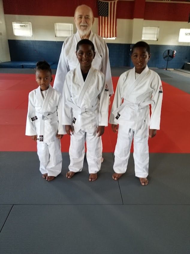 Three martial arts students in white gis with an instructor in a dojo on red and gray mats.