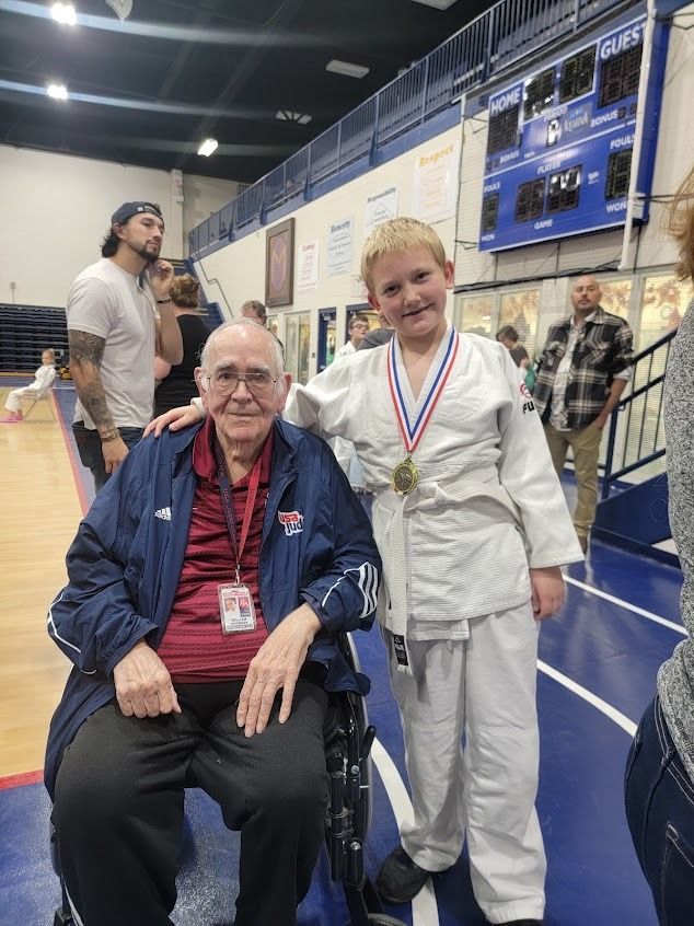 Two people posing indoors at a gym, one seated in a red-white-blue medal and white gi, one standing beside him.