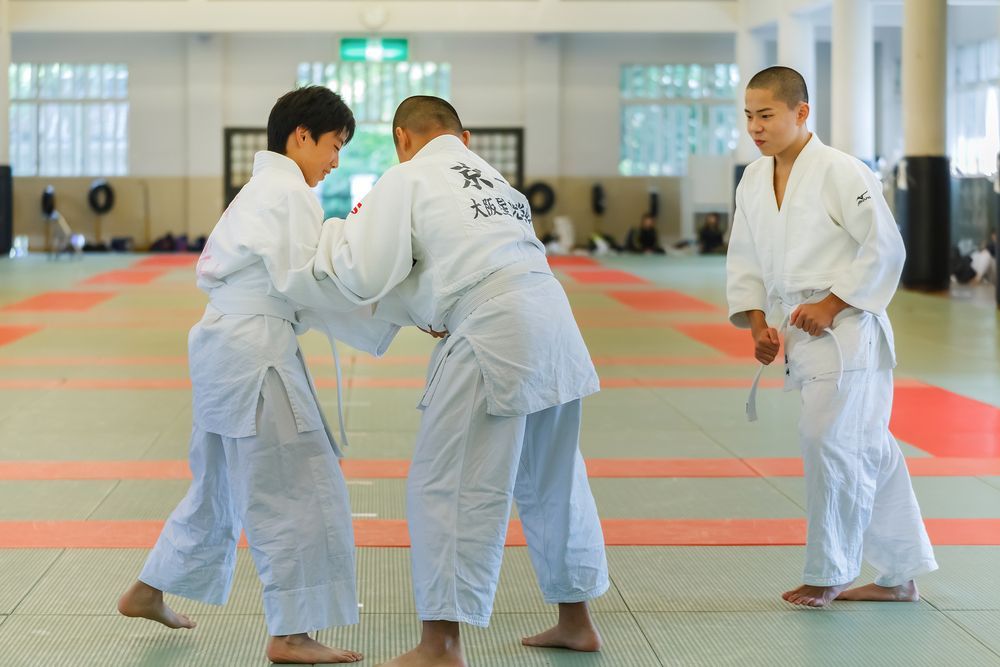 Three martial artists in white gis practice grappling on a dojo mat.