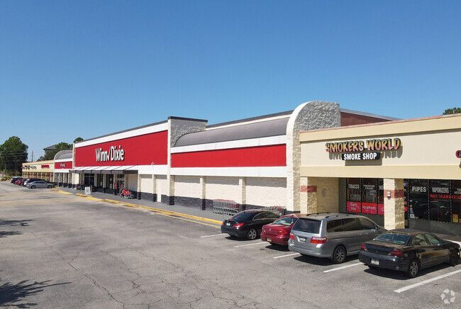 Exterior of a strip mall on a sunny day. Red signage on storefronts, cars parked out front.