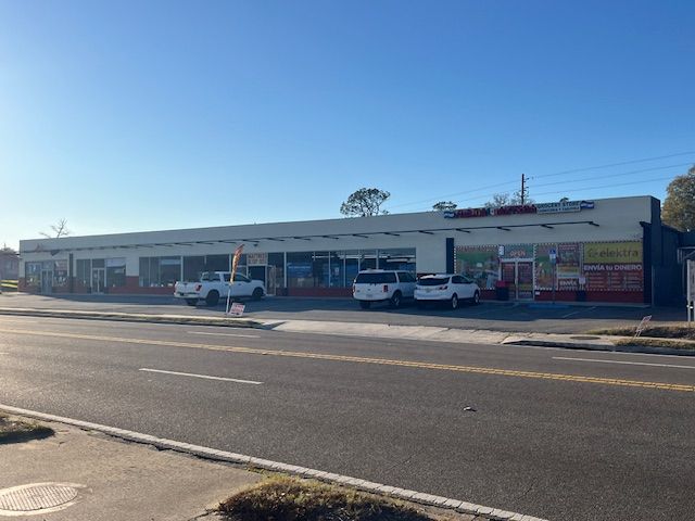 Shops in a strip mall on a sunny day. Cars are parked outside. A road is in the foreground.