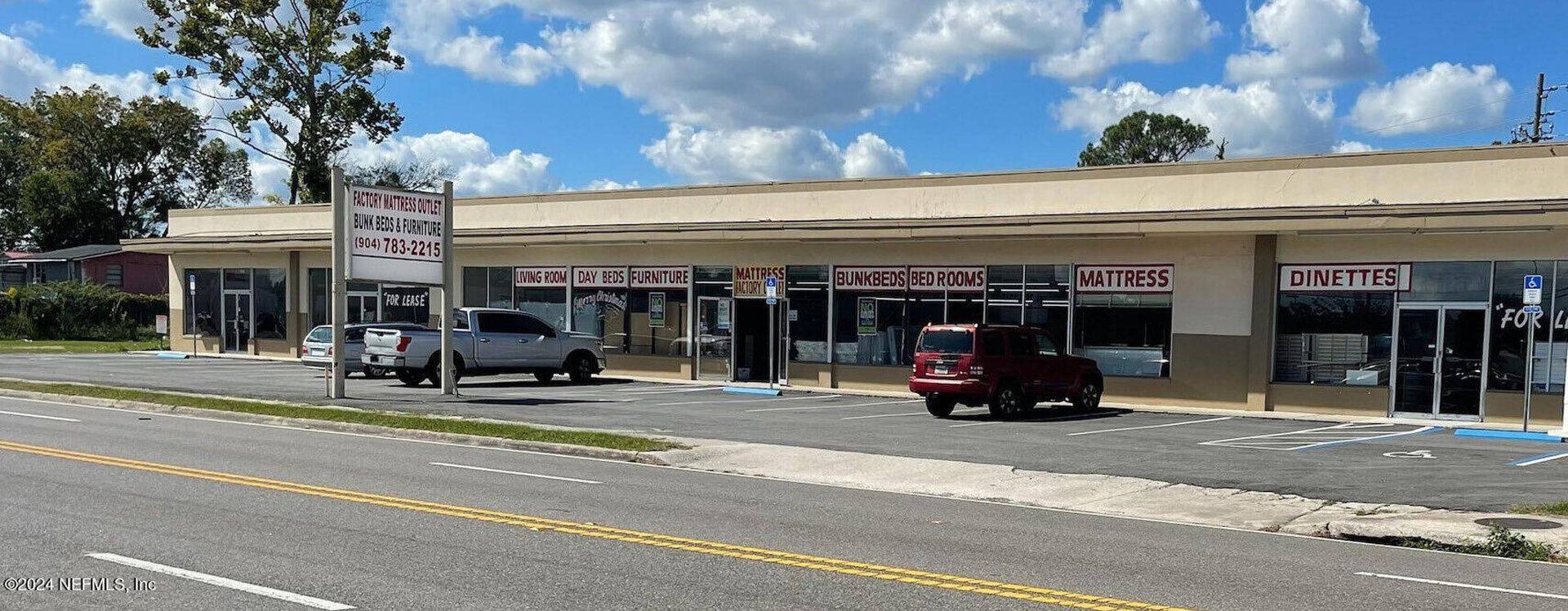 Shops in a strip mall on a sunny day. Cars are parked outside. A road is in the foreground.