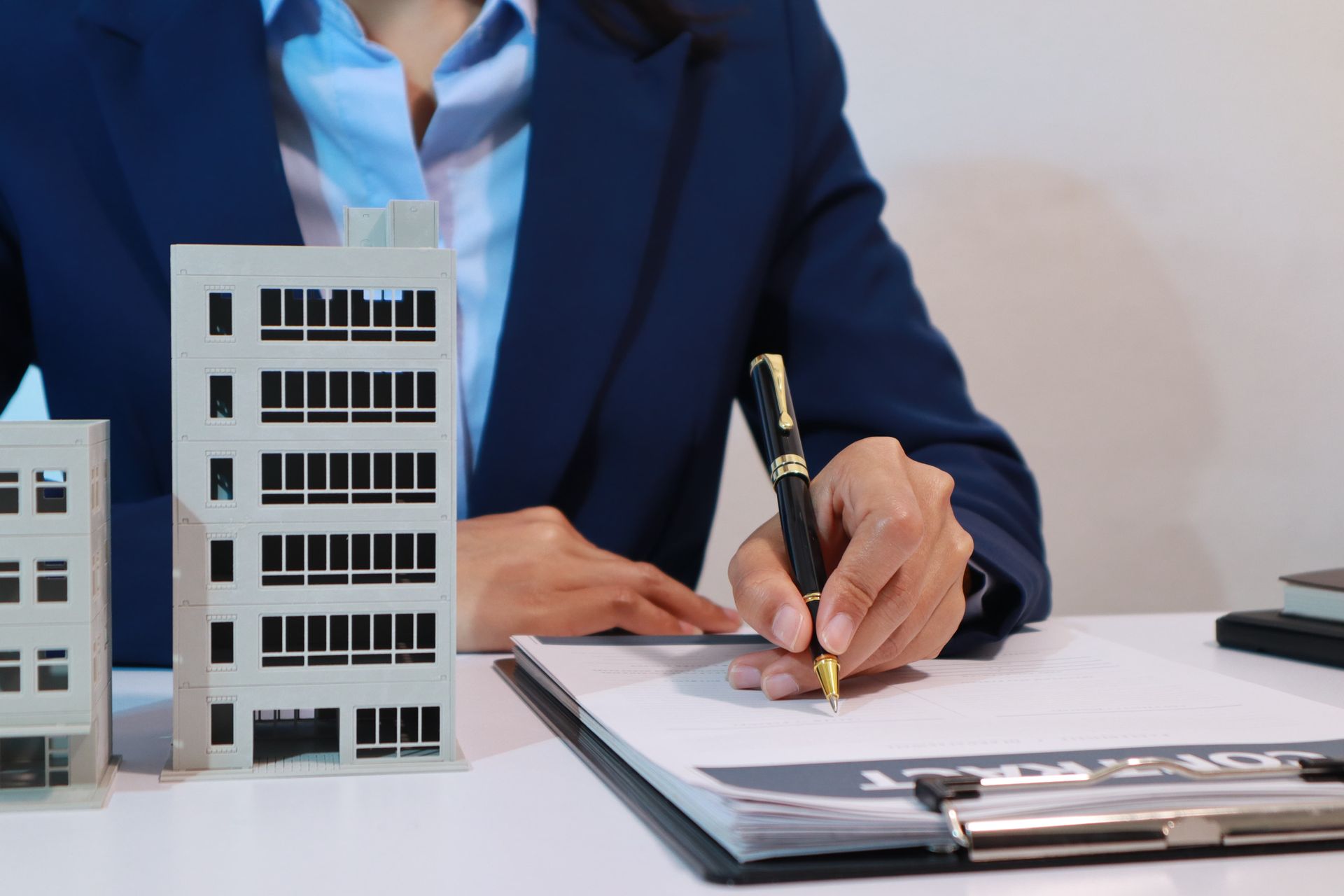 Person in suit signing paperwork next to building models, office setting.