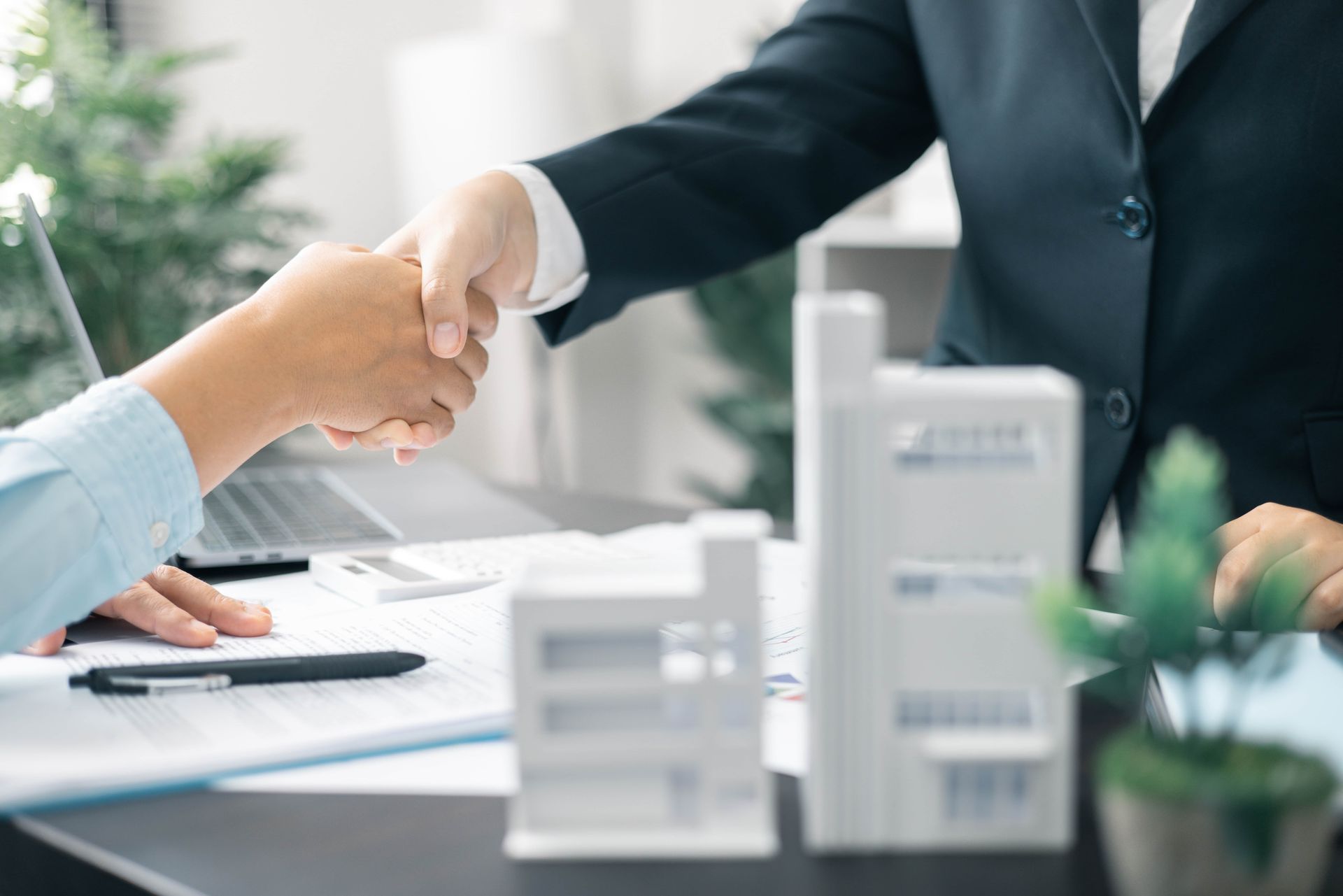 Two people shaking hands over a desk with building models and paperwork.
