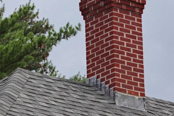 Red brick chimney rising from a gray shingled roof against a cloudy sky.
