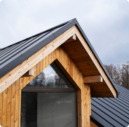Modern wooden house gable with dark metal roof and large triangular window under cloudy sky