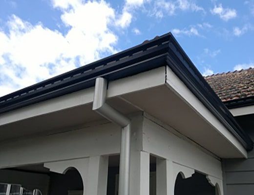 Close-up of a house roof corner with white guttering and dark trim against a blue sky.