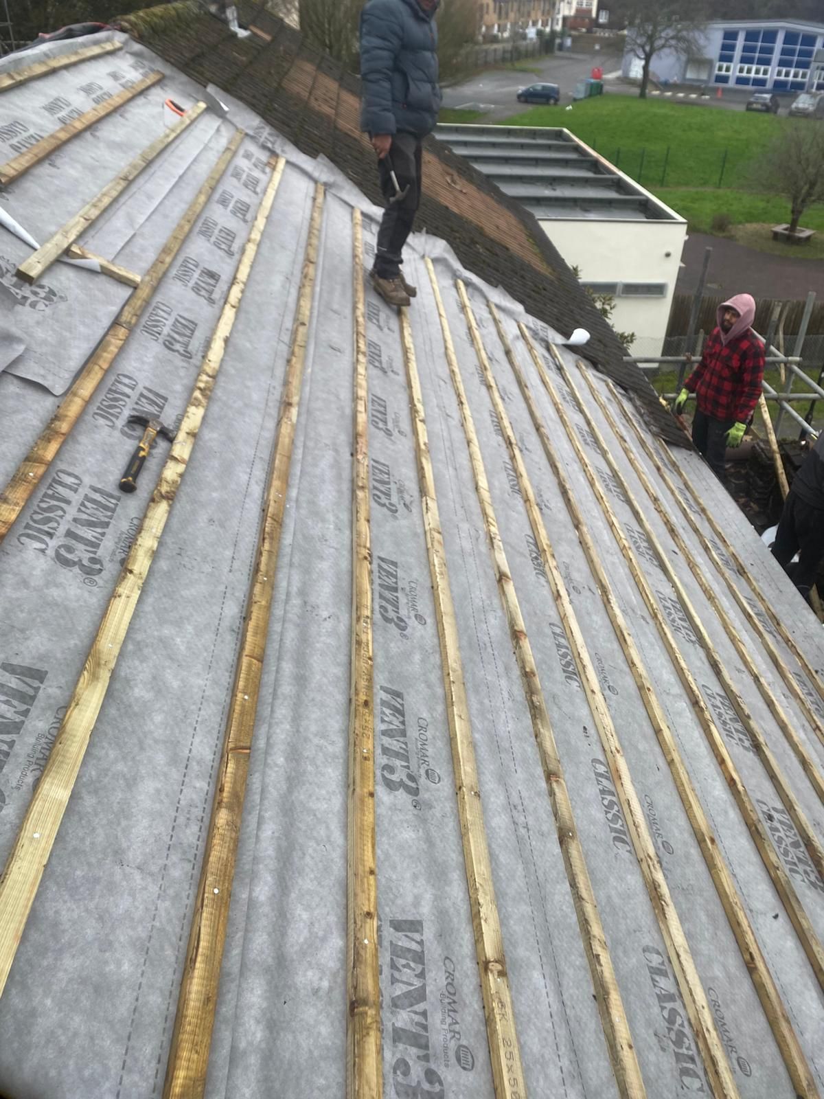 Workers on a steep roof installing silver insulation sheets with gold seams beside a grassy courtyard.