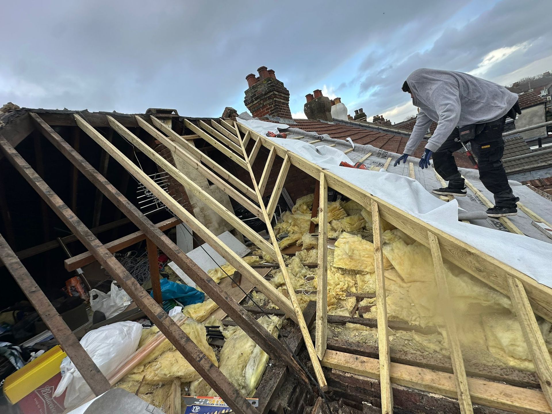 Worker installing roofing on a partially stripped house roof with exposed wooden rafters.