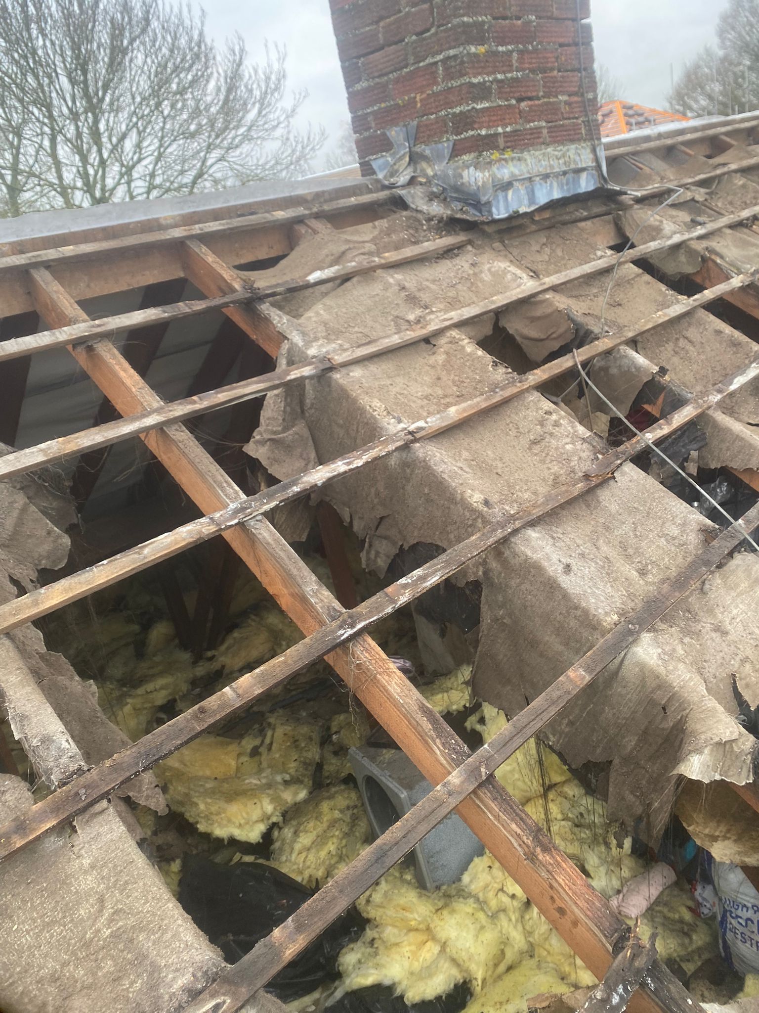 Damaged roof with missing tiles, exposed rafters, insulation, and a brick chimney.