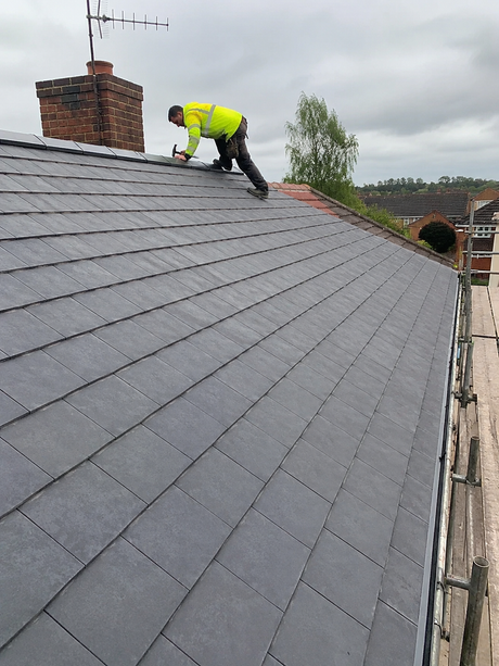 Worker in a hi-vis jacket inspecting a gray slate roof near a chimney.