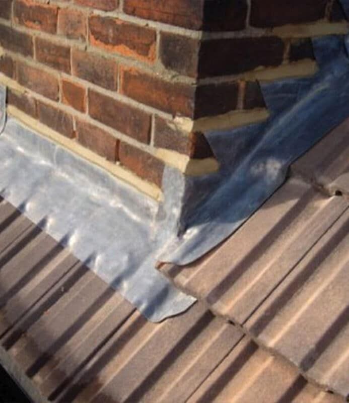 Brick chimney flashing on a corrugated metal roof, with blue sealant around the base.
