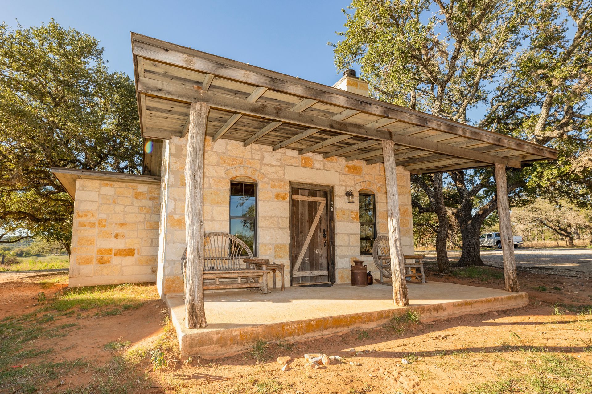 Small stone cabin with a porch, set in a grassy area with trees and a bright sky.