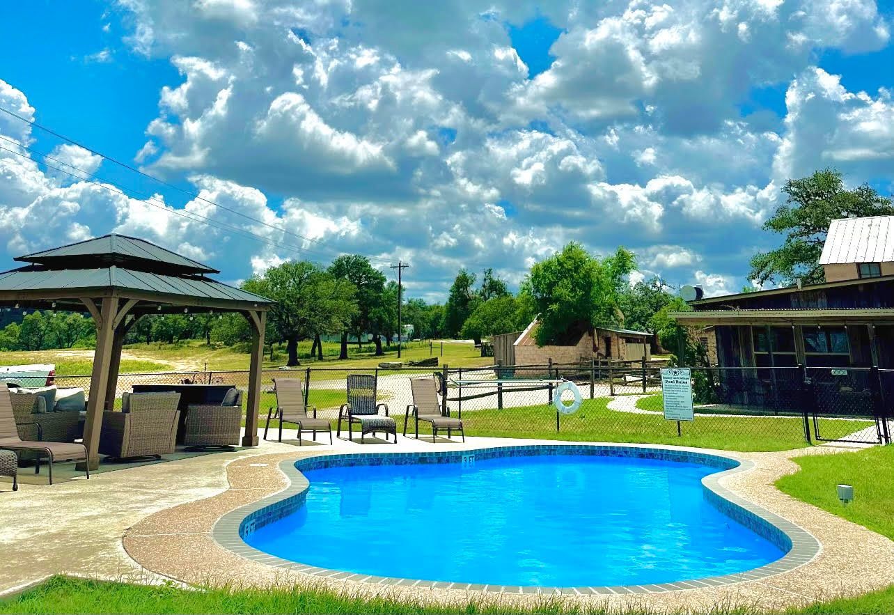 Pool with blue water, gazebo, chairs, and cloudy sky. Green grass, trees, and a building are in the background.