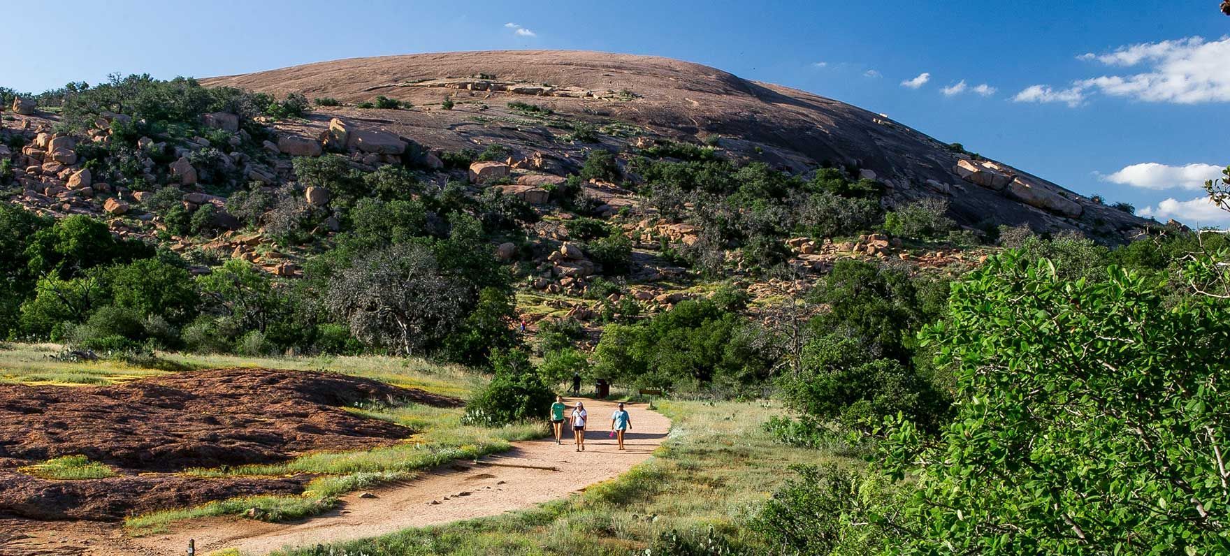 Hikers on a dirt trail with green shrubs below a rocky, sunlit hill under a blue sky